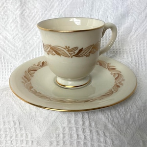 May include: Cream-colored teacup and saucer set with gold trim and a decorative leaf pattern. The teacup has a handle and sits on a small pedestal. The saucer is round and also features the leaf design. A classic tea set.