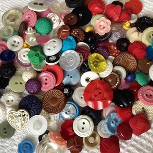 May include: A collection of various buttons in different colors, shapes, and sizes. The buttons include round, square, and flower-shaped designs. Colors range from white, pink, and red to green, blue, and black. Some buttons have intricate patterns and textures.