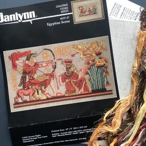May include: A Janlynn cross-stitch kit depicting an Egyptian scene. The kit includes pre-sorted floss, a needle, and instructions. The finished size is 38.1 cm x 22.9 cm. The design features figures in traditional Egyptian attire, with hieroglyphs and detailed imagery.