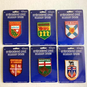 May include: Six embroidered crest patches on blue backing cards. Each patch features a provincial or territorial emblem, including Northwest Territories, Saskatchewan, Nova Scotia, Newfoundland & Labrador, Manitoba, and Yukon. The text "embroidered crest ecusson brodé" is printed at the top.