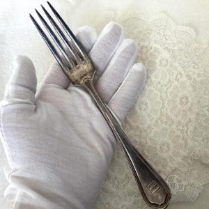 May include: A silver fork with four prongs, held in a white gloved hand. The fork has a decorative handle with a rectangular emblem near the end. The background includes white lace fabric.