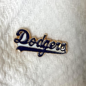 May include: A gold-toned metal lapel pin featuring the word Dodgers in a cursive script. The lettering is filled with dark blue enamel and accented with a white outline, creating a bold contrast against the metallic gold edges of the pin.