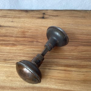 May include: A vintage metal door knob set featuring two rounded, dark-toned knobs connected by a central square spindle. The hardware displays a weathered, oxidised finish with visible signs of age, including dark brown and grey metallic patinas.