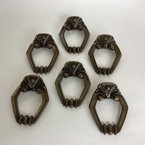 May include: Six antique brass cabinet door pulls with ornate designs. Each pull has a decorative ring and a central oval shape.