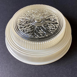 May include: A round ceiling light fixture cover featuring a clear glass centre with a pressed starburst and berry pattern. The glass is surrounded by a wide, ribbed, off-white plastic base with a stepped edge design and a beaded outer rim.