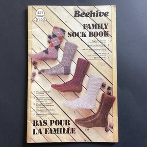 May include: A book titled "Beehive Family Sock Book" with a wooden background and five pairs of knitted socks in various colours and patterns. The book includes instructions for knitting argyle, two-colour, patterned, heel-less, and mesh socks.