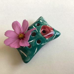 May include: A rectangular, teal ceramic flower frog with rounded corners and multiple circular openings. The surface features white painted streaks and a prominent red circular accent. A single pink cosmos flower with a yellow centre is inserted.