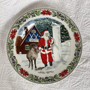 May include: A white ceramic plate featuring a Christmas scene. Santa Claus stands beside a reindeer in front of a red and blue building with a sign that reads "U.S. Post Office North Pole, N.Y.". The plate is decorated with a green wreath and red poinsettia flowers.