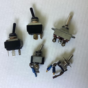 May include: A collection of five electrical toggle switches with various metal and black plastic housings. Some switches feature black levers while others have silver-toned metal levers. Several units include attached wire connectors and screw terminals.