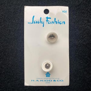 May include: Vintage card of two buttons. The card is white with the text "Lady Fashion" in blue script. The buttons are round, one with a tree design and the other with a circular pattern. The card also has the text "CANADA'S LARGEST BUTTON HOUSE".