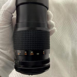 May include: A black camera lens with a textured grip and distance markings. The lens has a series of numbers indicating focal lengths, including 30, 50, and 100, and a measurement scale in feet.