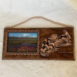 May include: A wooden plaque with a picture of Denali National Park in Alaska. The plaque has a copper-colored frame and a copper-colored relief sculpture of a dog sledding team. The text "Denali National Park Alaska" is printed on the plaque.