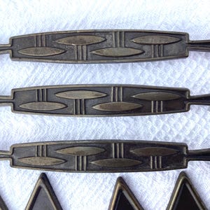 May include: Three horizontal metal drawer pulls with a dark bronze finish, featuring a repeating geometric pattern of elongated oval shapes and vertical lines. Each pull has a central rectangular body with tapered ends and triangular mounting points.