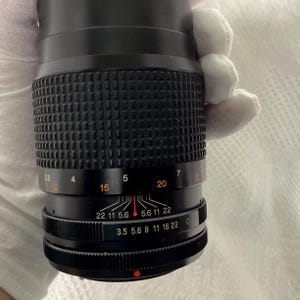 May include: A black camera lens with a ridged grip and various numerical markings. The lens has a focus ring with numbers from 3.5 to 22 and a distance scale. The lens is held by a gloved hand.