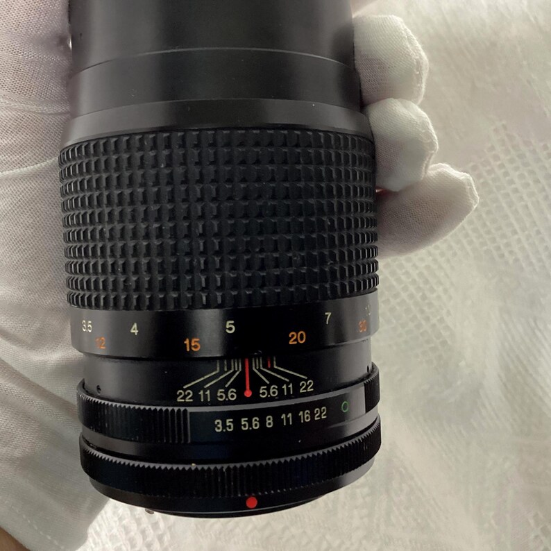 May include: A black camera lens with a ridged grip and various numerical markings. The lens has a focus ring with numbers from 3.5 to 22 and a distance scale. The lens is held by a gloved hand.