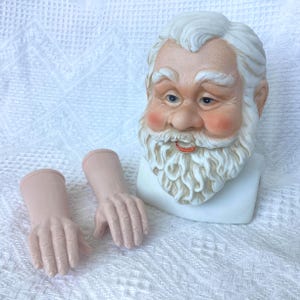May include: A ceramic Santa Claus bust with a white beard, rosy cheeks, and blue eyes. Two detached, pale pink ceramic hands are positioned in front of the bust. The bust is on a white surface.