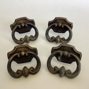 May include: Four antique brass cabinet drawer pulls with a textured finish and ornate design. Each pull has a ring handle and a decorative centre piece.