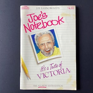 May include: A vintage paperback book titled "Joe's Notebook" with a portrait of a smiling man. The cover features pink and purple text, with the subtitle "It's a Taste of Victoria." A yellow pencil rests on the cover.