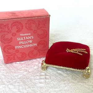 Peut inclure: Un coussin à épingles Sultan's Pillow vintage de Watkins. Le coussin en velours rouge repose sur une base en métal doré. La boîte rose porte le nom du produit. Une chaîne dorée est attachée au coussin.
