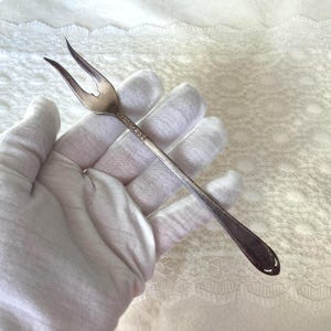 May include: A silver-toned metal cocktail fork featuring two slender, curved tines and a decorative embossed pattern along the upper handle. The handle tapers towards a rounded, smooth end, showcasing a classic metallic finish and intricate detailing.