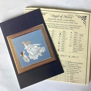 May include: A framed cross-stitch design titled "Angel of Mercy" featuring an angel figure in white and gold, holding a smaller figure. The artwork is set against a light blue background, with a gold-colored frame. The design is on a dark blue textured surface, with a pattern chart.
