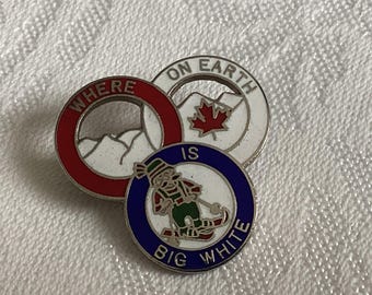 May include: A silver pin with three interlocking circles. The top circle is white with red lettering that says "Where on Earth is Big White". The middle circle is red with white lettering that says "Where". The bottom circle is blue with white lettering that says "Big White". A cartoon skier is in the center of the bottom circle.