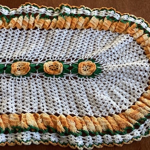 May include: White crocheted oval table runner with a green and orange border. The border features a scalloped edge and a row of crocheted flowers.