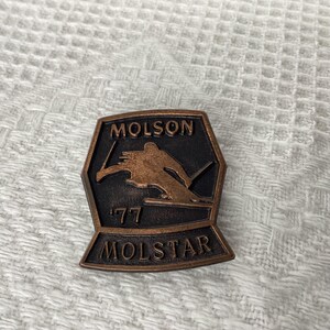 May include: A bronze-colored metal pin with a skier in silhouette, the text "Molson" and "Molstar" and the number "'77".