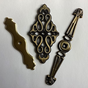May include: Three decorative metal hardware pieces in antique gold and black. The pieces include a curved handle, an ornate diamond-shaped accent, and a pull with a circular centre. These are likely cabinet or furniture hardware.