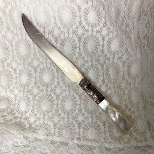 May include: A vintage butter knife with a silver blade and a decorative handle. The handle features a white, pearlescent grip and ornate silver detailing. The knife is approximately 20 cm long.