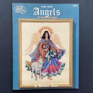 May include: Cross stitch pattern titled "Angels" featuring six heavenly designs. The cover shows a framed cross-stitch image of an angel with a wolf. The angel is wearing a colorful robe and has large wings. The text "American School of Needlework" is also visible.