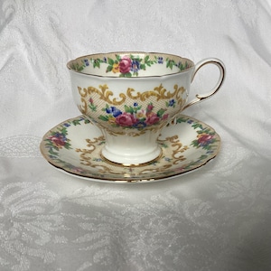 May include: A vintage teacup and saucer set with a floral pattern and gold accents. The cup and saucer are white with a beige background and feature a delicate floral design with pink, blue, and yellow flowers. The gold accents are around the rim of the cup and saucer and on the handle of the cup.