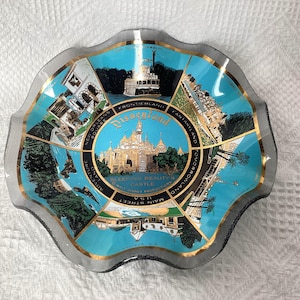 May include: A clear glass dish with a scalloped edge, featuring a teal and gold design with images of Disneyland attractions. The center displays Sleeping Beauty's Castle, surrounded by text and illustrations of various park areas.