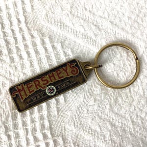 May include: A rectangular Hershey's keychain with a gold-colored key ring. The keychain has a brown background with the word "HERSHEY'S" in red letters. The text "EST 1894" is also visible.
