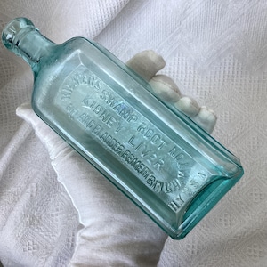 May include: A rectangular, light aqua-coloured glass bottle with embossed text reading KILMER'S SWAMP ROOT NO. 1 KIDNEY LIVER AND BLADDER REMEDY BINGHAMTON NY USA. The bottle features a narrow neck and a rounded rim, showcasing a vintage medicinal design.
