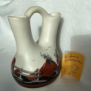 May include: A white ceramic wedding vase with a brown and black painted design of a landscape with mountains, birds, and a fence. The vase has two spouts and a handle. The vase is accompanied by a yellow tag that reads "The Legend of the Wedding Vase".