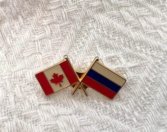May include: A gold-toned pin featuring two crossed flags. The flag on the left is the Canadian flag, with a red maple leaf on a white background. The flag on the right is the Russian flag, with white, blue, and red stripes.