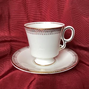 Peut inclure: Service à thé blanc avec des bordures dorées et un motif géométrique. La tasse à thé a un piedestal et une anse incurvée. La soucoupe présente un motif assorti sur le bord. L'ensemble est sur un fond rouge.
