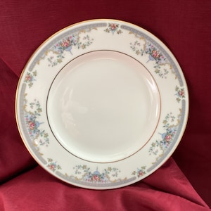 May include: A white ceramic plate with a gold rim and a floral pattern in shades of blue, pink, and green. The plate has a delicate, vintage aesthetic.
