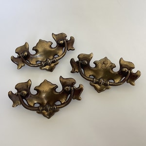 May include: Three antique brass drawer pulls with ornate, leaf-shaped designs. Each pull has a simple, curved handle.