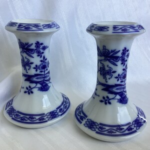 May include: A pair of white porcelain candle holders with a blue and white floral pattern. The holders have a flared base and a cylindrical top.