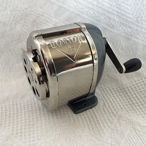 May include: A vintage Boston KS pencil sharpener featuring a polished chrome metal front plate with the embossed text BOSTON KS. The body is constructed from dark gray metal with a side-mounted hand crank handle featuring a black plastic knob.