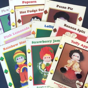 May include: A collection of vintage crochet pattern booklets with titles like "Popcorn," "Strawberry Jam," and "Taffy Apple." Each booklet features a colorful illustration of a crocheted character or food item, with cupcake icons.