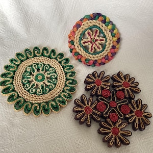 May include: Three decorative woven coasters in various designs. One coaster is circular with green and beige accents. Another is a colourful braided circle. The third set features flower-shaped coasters with red and brown hues.