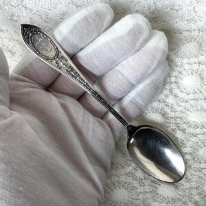 May include: A silver-toned metal spoon featuring an ornate, embossed handle with intricate scrollwork and a central oval cartouche containing a decorative monogram. The spoon has a smooth, polished, oval-shaped bowl and a tapered, detailed stem.