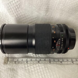 May include: A black camera zoom lens with measurement markings. The lens has a ribbed grip section and various numerical settings. A ruler is placed next to the lens, showing measurements in inches.
