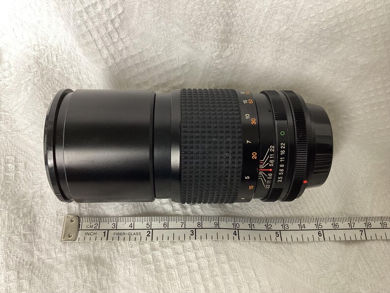 May include: A black camera zoom lens with measurement markings. The lens has a ribbed grip section and various numerical settings. A ruler is placed next to the lens, showing measurements in inches.