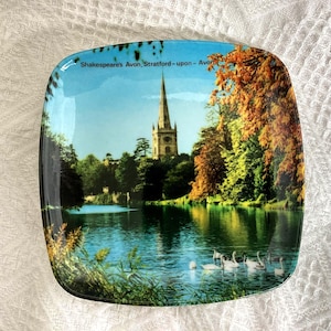 May include: Square decorative plate with a scenic image of a lake, swans, trees, and a church spire. The plate has a glossy finish and the text "Shakespeare's Avon, Stratford-upon-Avon". The image has a vintage aesthetic.