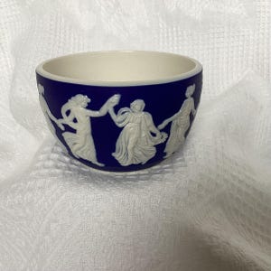 May include: A small, round, blue and white ceramic bowl. The bowl's exterior is a deep blue, decorated with white bas-relief figures in a classical style. The interior is a creamy white colour. The bowl is likely for decorative use.