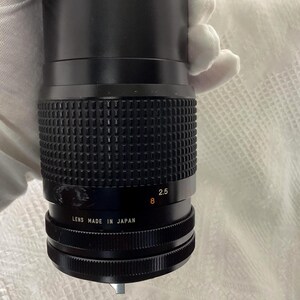 May include: Black camera lens with a ridged grip and a black barrel. The lens has white text that reads "2.5" and "8". The bottom of the lens reads "LENS MADE IN JAPAN".